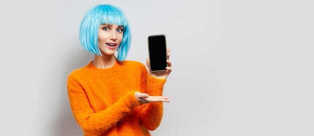 Studioporträt des jungen blauhaarigen Mädchens, das ein Smartphone hält, auf grauer Wand.