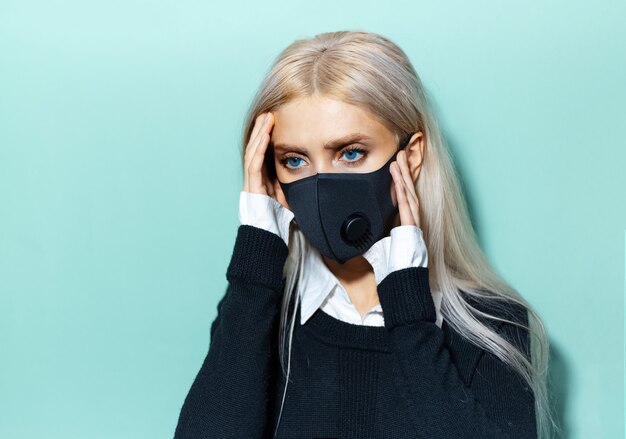 Studioporträt des jungen blonden Mädchens, das Kopfschmerzen trägt, die schwarze medizinische Maske auf Cyan tragen