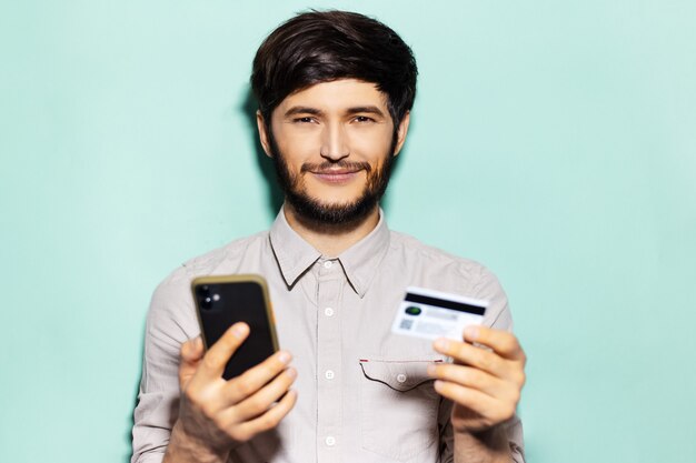 Studioporträt des jungen positiven Kerls, der Kamera betrachtet, unter Verwendung des Smartphones und der Kreditkarte auf Hintergrund der aqua menthe Farbe.