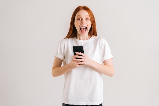 Studioporträt einer fröhlichen jungen Frau mit offenem Mund, die eine gute Online-Nachricht mit dem Handy liest