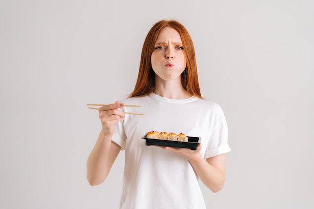 Studioporträt einer hungrigen jungen Frau, die köstliche Sushi-Rollen mit Essstäbchen isst und in die Kamera blickt, die auf weißem, isoliertem Hintergrund steht. Hübsche kaukasische Rothaarigefrau, die asiatische Nahrung kaut.