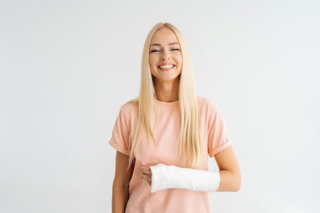 Studioporträt einer positiven blonden jungen Frau mit gebrochenem Arm, eingewickelt in Gipsbandage, die lächelnd in die Kamera blickt, die auf weißem, isoliertem Hintergrund steht