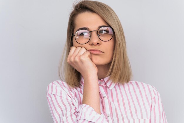 Studioporträt einer traurigen blonden Studentin mit rosafarbenem Hemd und runder Brille, die mit unglücklichem, langweiligem Ausdruck auf ihrem Gesicht sitzt und das Kinn auf der Hand ruht, um Informationen zu hören