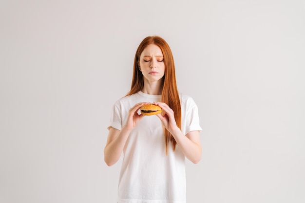 Studioporträt einer unzufriedenen hübschen jungen Frau mit Ekel, die einen Burger hält und nach unten auf weißen, isolierten Hintergrund blickt Traurige süße rothaarige Frau, die Hamburger isst