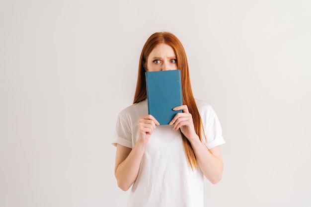 Studioporträt einer verängstigten jungen Studentin versteckt das Gesicht hinter einem Notizbuch, das in die Kamera blickt und auf weißem, isoliertem Hintergrund steht. Erstaunte rothaarige Dame, die mit Lehrmaterialien aufwirft.