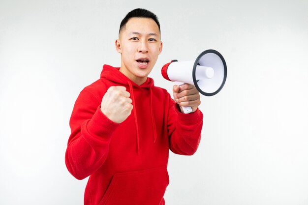 Studioporträt eines jungen asiatischen Mannes in einem roten Kapuzenpulli mit einem Lautsprecher in seinen Händen auf einem weißen Hintergrundstudio.