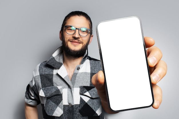 Studioporträt eines jungen lächelnden Mannes, der ein großes Smartphone mit leerem Bildschirm in der Hand hält und in der Nähe der Kamera ein Gerät mit Mockup auf weißem Hintergrund zeigt, das ein grau kariertes T-Shirt und eine Brille trägt