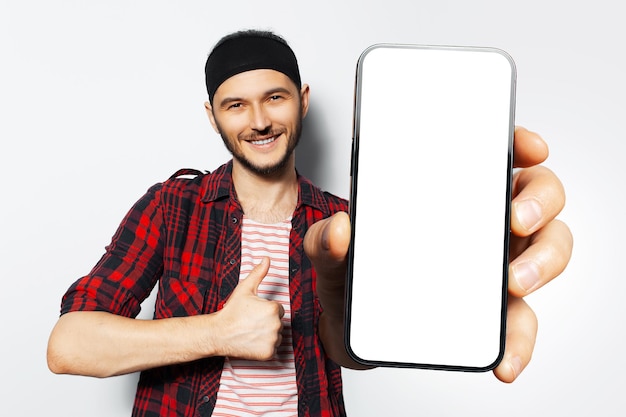 Studioporträt eines jungen lächelnden mannes, der ein großes smartphone mit leerem bildschirm in der hand hält und in der nähe der kamera ein gerät mit mockup und daumen nach oben auf weißem hintergrund zeigt