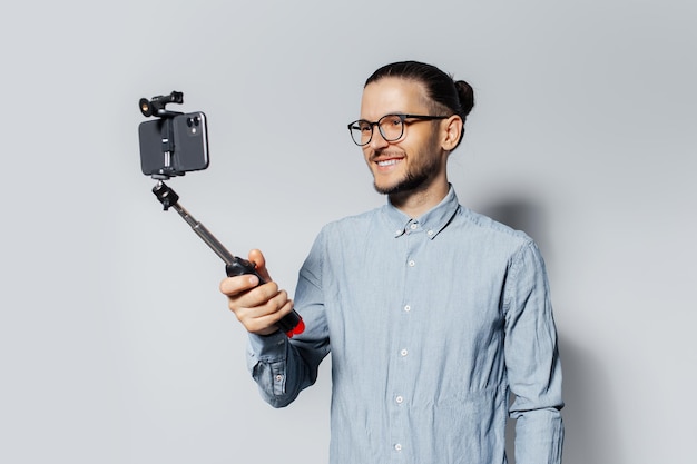 Studioporträt eines jungen lächelnden Mannes, der ein Selfie-Foto per Smartphone macht und auf weißem Hintergrund haftet. Mit Brille und blauem T-Shirt