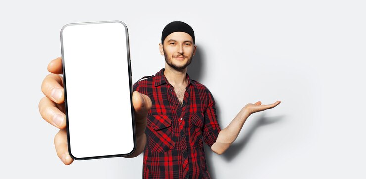 Studioporträt eines jungen lächelnden Mannes, der in der Hand ein großes Smartphone mit leerem Bildschirm in der Hand hält und in der Nähe der Kamera ein Gerät mit Mockup-Punkthand zeigt, das mit einer Handfläche auf Weiß präsentiert oder demonstriert
