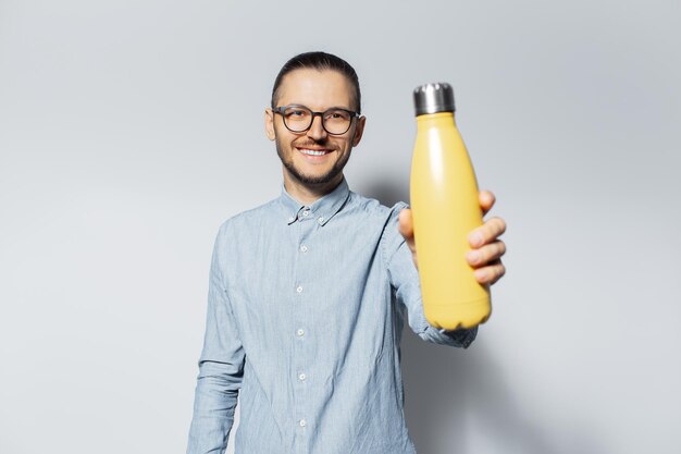 Studioporträt eines jungen lächelnden Mannes, der in der Nähe der Kamera eine gelbe Thermowasserflasche aus Stahl auf hellgrauem Hintergrund zeigt