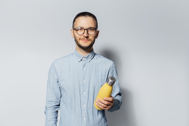 Studioporträt eines jungen Mannes, der eine gelbe Thermowasserflasche aus Stahl in den Händen auf hellgrauem Hintergrund hält Mit Brille und T-Shirt