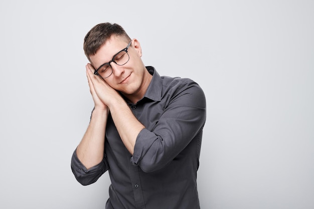 Studioporträt eines jungen Mannes mit Brille und Hemd, der Handflächen unter den Kopf legt und so tut, als würde er isoliert auf weißem Hintergrund schlafen