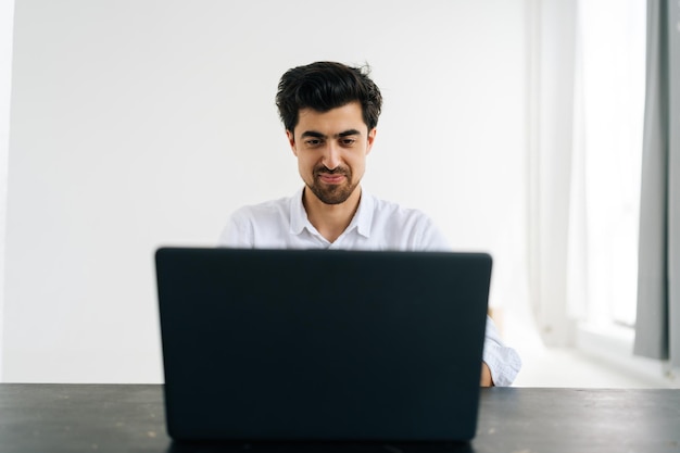 Studioporträt eines lächelnden bärtigen Mannes im Hemd, der auf einem Laptop-Computer tippt und auf den Bildschirm schaut, der am Schreibtisch in einem Zimmer mit hellem Interieur sitzt