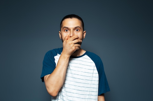 Studioporträt eines Mannes, der den Mund mit der Hand auf blauem Hintergrund bedeckt T-Shirt trägt