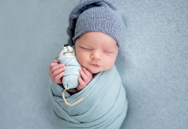 Studioportrait des neugeborenen Babys mit Spielzeugmaus. Entzückendes Säuglingskind, das mit Hut schläft und in Stoff gewickelt ist