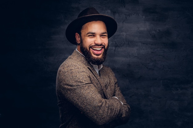 Studioportrait eines lächelnden hübschen afroamerikanischen Mannes in einer eleganten braunen Jacke und einem Hut. Getrennt auf einem dunklen Hintergrund.