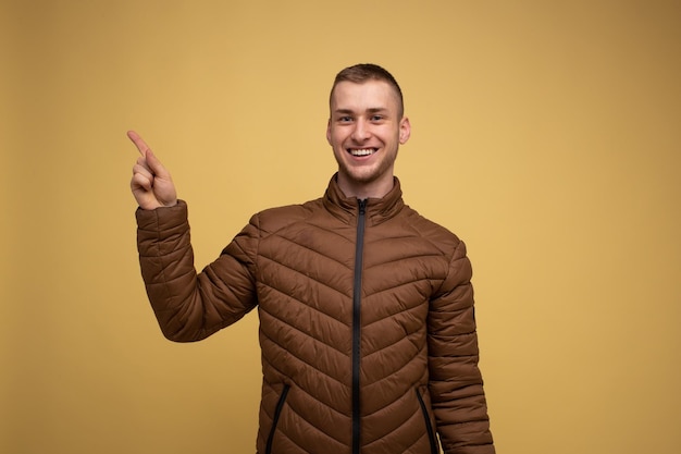 Studioportrait Junger 20er Mann in brauner Jacke auf gelbem Hintergrund lacht und zeigt mit den Fingern in die Richtung