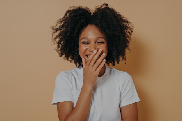 Studioportrait von glücklichem biracial Mädchen hautnah. Schöne dunkelhäutige tausendjährige Frau, die sich beim Lachen den Mund mit der Hand bedeckt. Nettes positives Mischrennen-Weibchen lokalisiert über beigem Hintergrund