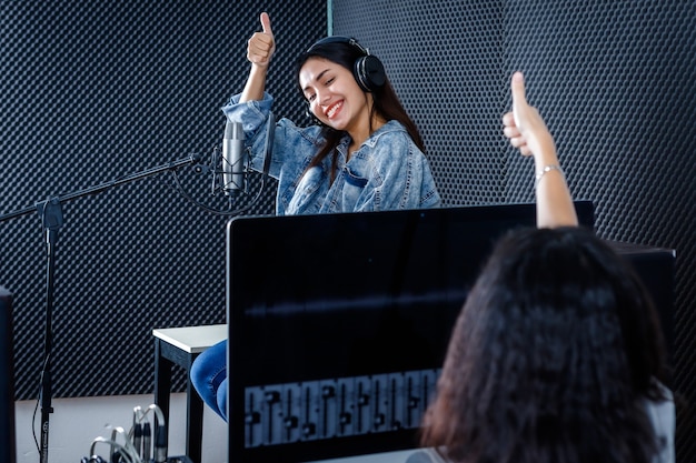 Studiotechnikerin des Computermonitors der Software für die Aufnahme im Vordergrund der jungen asiatischen Sängerin mit Kopfhörern, die in einem professionellen Studio ein Lied vor dem Mikrofon aufnimmt