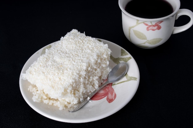 Stück Couscous in Porzellan Untertasse auf schwarzem Hintergrund Typisches brasilianisches Gericht mit Tapioka-Milchzucker und Kokosmilch Mit Tasse Kaffee