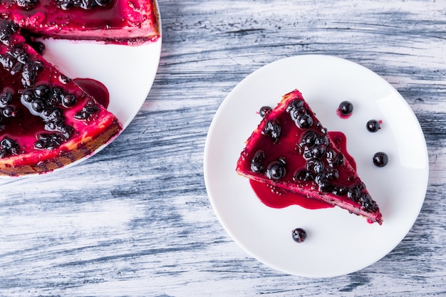 Stück Käsekuchen auf weißem Teller mit Johannisbeere auf farbigem Holzhintergrund. Draufsicht.