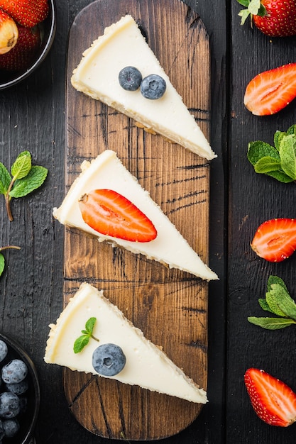 Stück Käsekuchen mit Erdbeeren, Blaubeeren und Minze, auf schwarzem Holztischhintergrund, Draufsicht flach