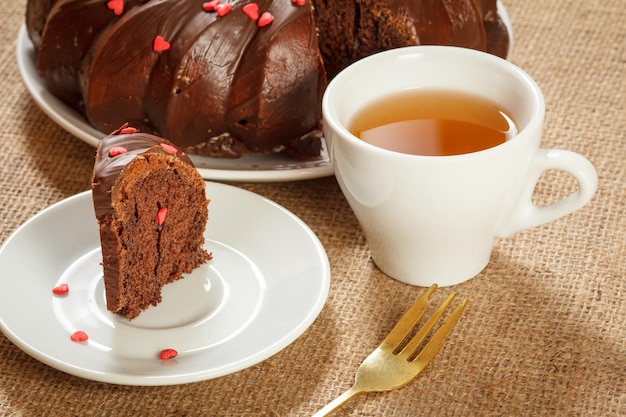 Stück köstlicher hausgemachter Schokoladenkuchen, verziert mit kleinen Herzen aus Karamell auf Untertasse, Gabel und einer Tasse Tee auf dem Tisch mit Sackleinen.