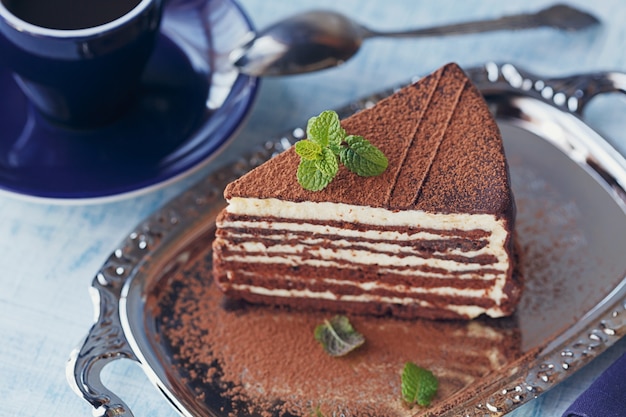 Stück köstlicher Schokoladenkuchen auf silbernem Teller mit frischer Minze auf hellblauem Holztischhintergrund. Nahaufnahme.