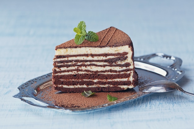 Stück köstlicher Schokoladenkuchen auf silbernem Teller mit frischer Minze auf hellblauem Holztischhintergrund.