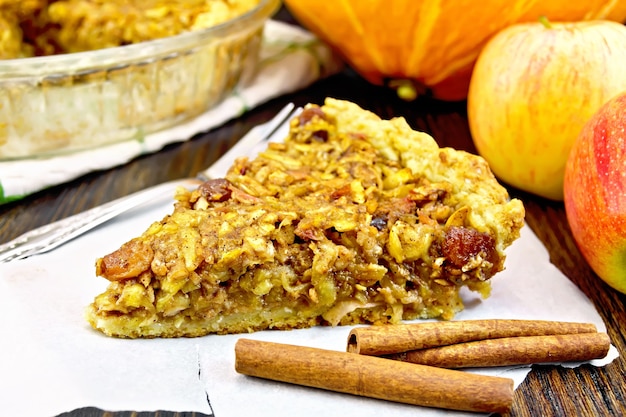 Stück Kuchen mit Äpfeln, Kürbis, Rosinen und Nüssen auf Pergament, Zimt auf dem Hintergrund eines Holzbrettes