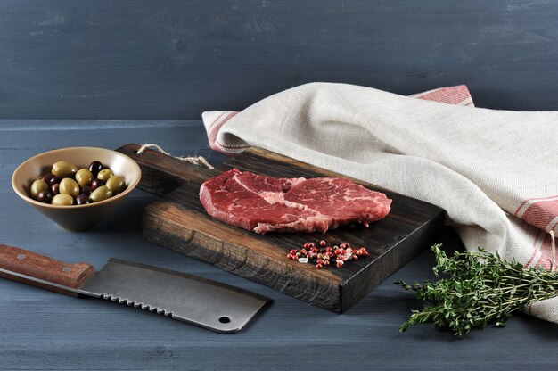 Stück rohes Rindfleisch auf einem Holzbrett, ein Messer für Fleisch, Oliven und Thymian