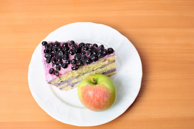 Stück süßer Heidelbeerkuchen mit violetter Sahne und grünem Apfeldessert auf weißer Platte, die auf Holztischhintergrund steht