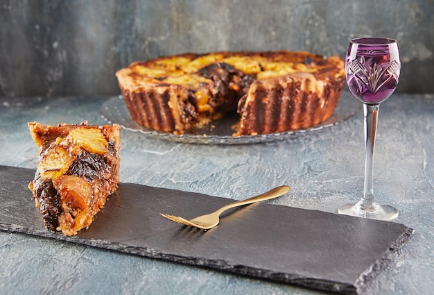 Stück winterkuchen mit aprikosen, pflaumen, bananen und ananas, auf schwarzem teller mit schuss likör und gabel. französische gourmetküche