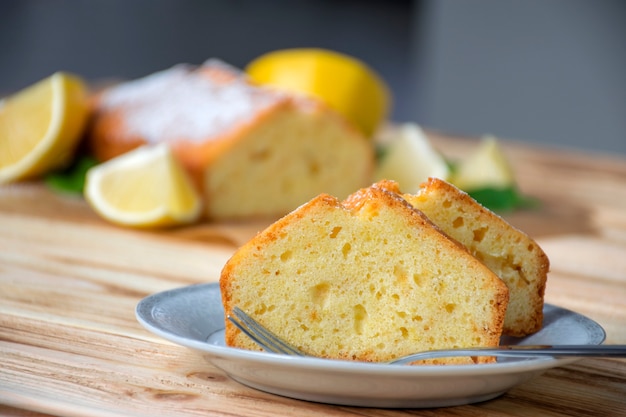 Stück Zitronenkuchen auf Teller auf rustikalem Holzbrett mit vollem Kuchen und Zitronen auf Hintergrund.