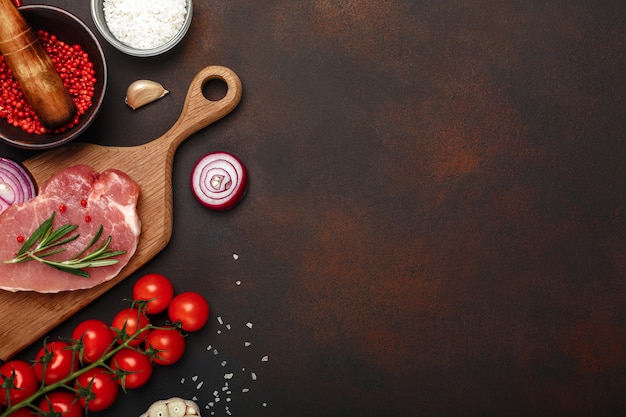 Stücke rohes Schweinefleischsteak auf Schneidebrett mit Kirschtomaten, Rosmarin, Knoblauch, rotem Pfeffer, Zwiebel, Salz