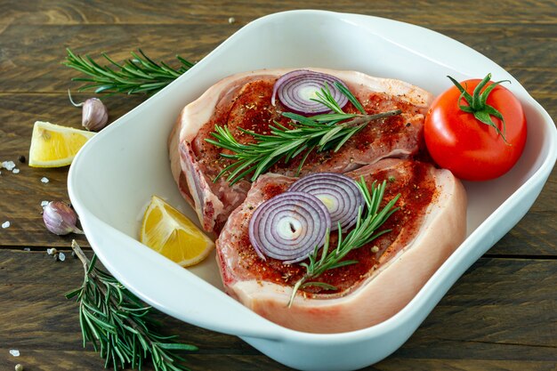 Stücke rohes Schweinesteak mit Gewürzen und Kräutern Rosmarin, Salz und Pfeffer in Keramikbackform auf Holzhintergrund im rustikalen Stil.