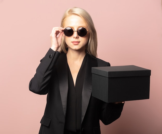 Style Blondine in Blazer und Sonnenbrille mit schwarzer Geschenkbox auf Pink