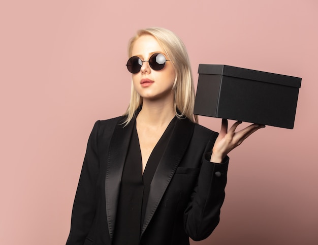 Style Blondine in Blazer und Sonnenbrille mit schwarzer Geschenkbox auf Pink