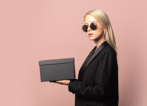 Style Blondine in Blazer und Sonnenbrille mit schwarzer Geschenkbox auf Pink