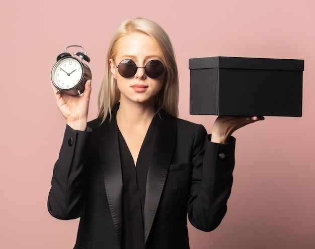 Style Blondine in Blazer und Sonnenbrille mit schwarzer Geschenkbox und Wecker auf Pink