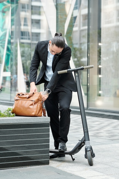 Stylidh junger Unternehmer mit Roller auf der Suche nach Dokument in Ledertasche beim Telefonieren mit Kollegen