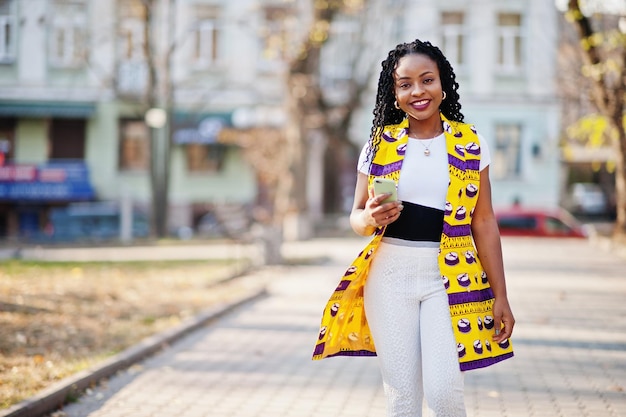 Stylische afroamerikanische Frauen in gelber Jacke posierten an sonnigen Tagen mit dem Handy zur Hand auf der Straße