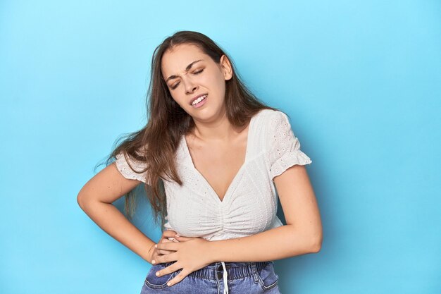 Stylische junge Frau in weißer Bluse auf einem blauen Studio-Hintergrund mit Leber- und Magenschmerzen