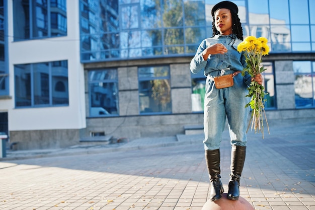 Stylische, modische afroamerikanische Frauen in Jeans und schwarzer Baskenmütze mit gelbem Blumenstrauß posierten im Freien an sonnigen Tagen vor blauem, modernem Gebäude