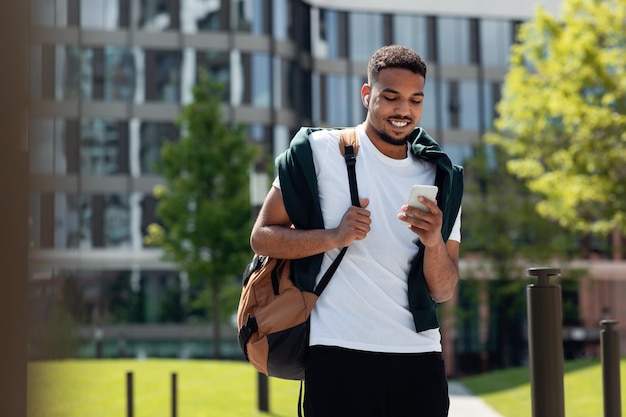 Stylischer Afroamerikaner mit drahtlosen Kopfhörern, der beim Gehen in der Stadt auf dem Smartphone SMS schreibt