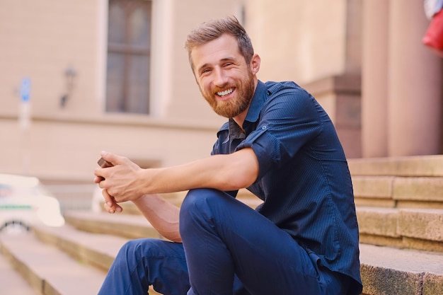 Stylischer bärtiger positiver Mann in Jeans und Jeansjacke sitzt auf einer Stufe und benutzt ein Smartphone.