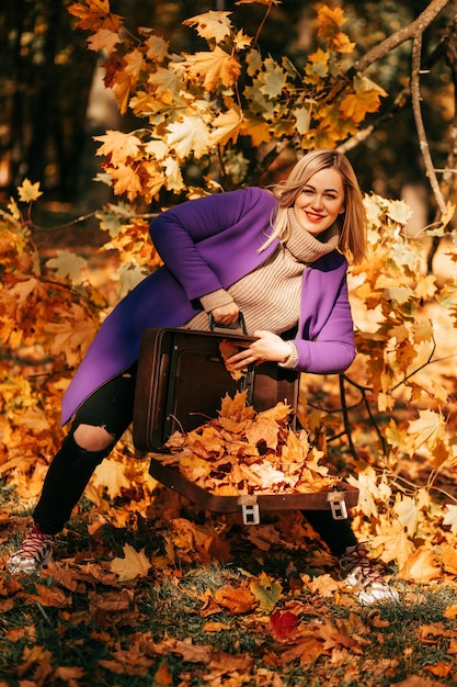 Stylischer blonder gestrickter Pullover und heller lila Mantel lächelt und zeigt einen Haufen gefallenen orangefarbenen Herbstes