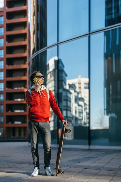 Stylischer Hipster mit Sonnenbrille hält Longboard in der Hand und spricht per Handy Freizeitbeschäftigung