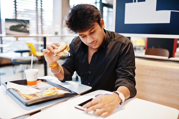 Stylischer indischer Mann, der im Fast-Food-Café sitzt und Hamburger isst, liest Morgennachrichten auf dem Handy